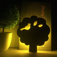 OptiLair tree-shaped acrylic night light with warm yellow glow and sound-sensor activation in a dark bedroom setting.